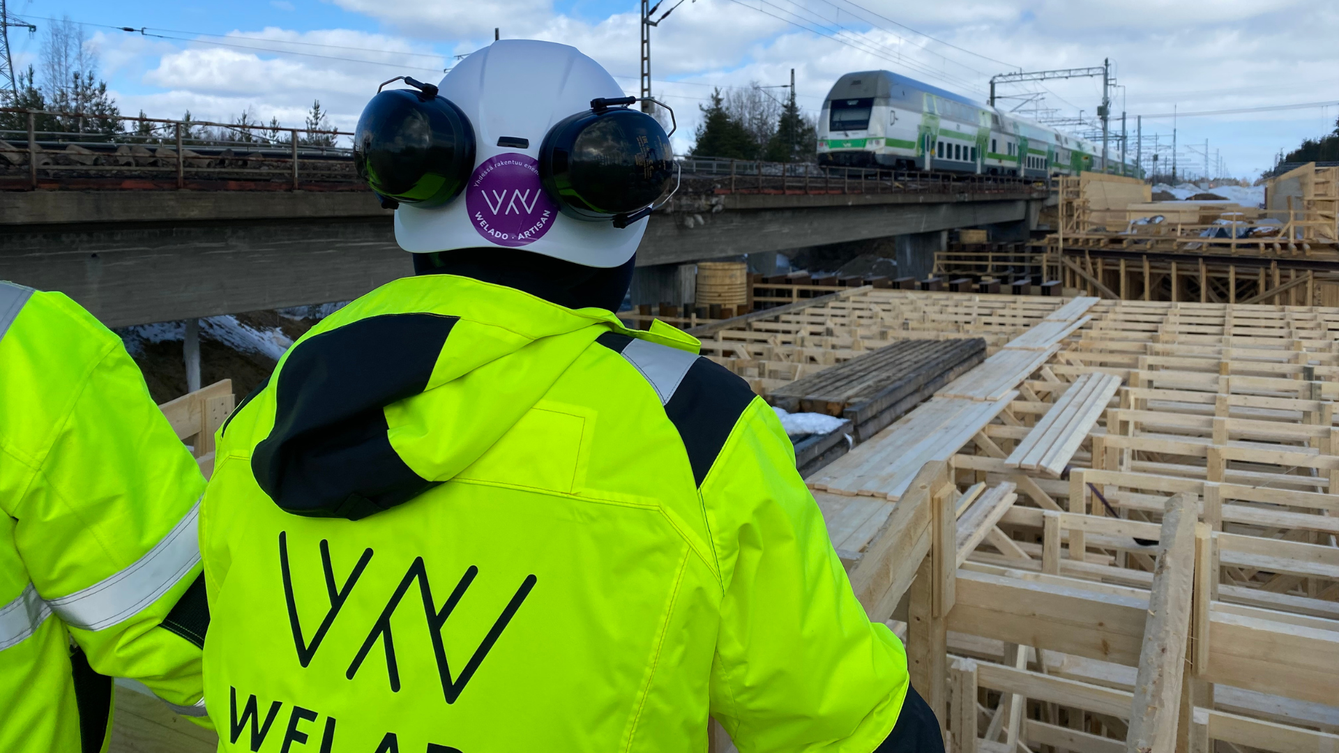 Weladolaisia junaradan varressa katselemassa rakennustyömaata neonvihreät huomiotakit päällä ja valkoset Weladon suojakypärät päässä.